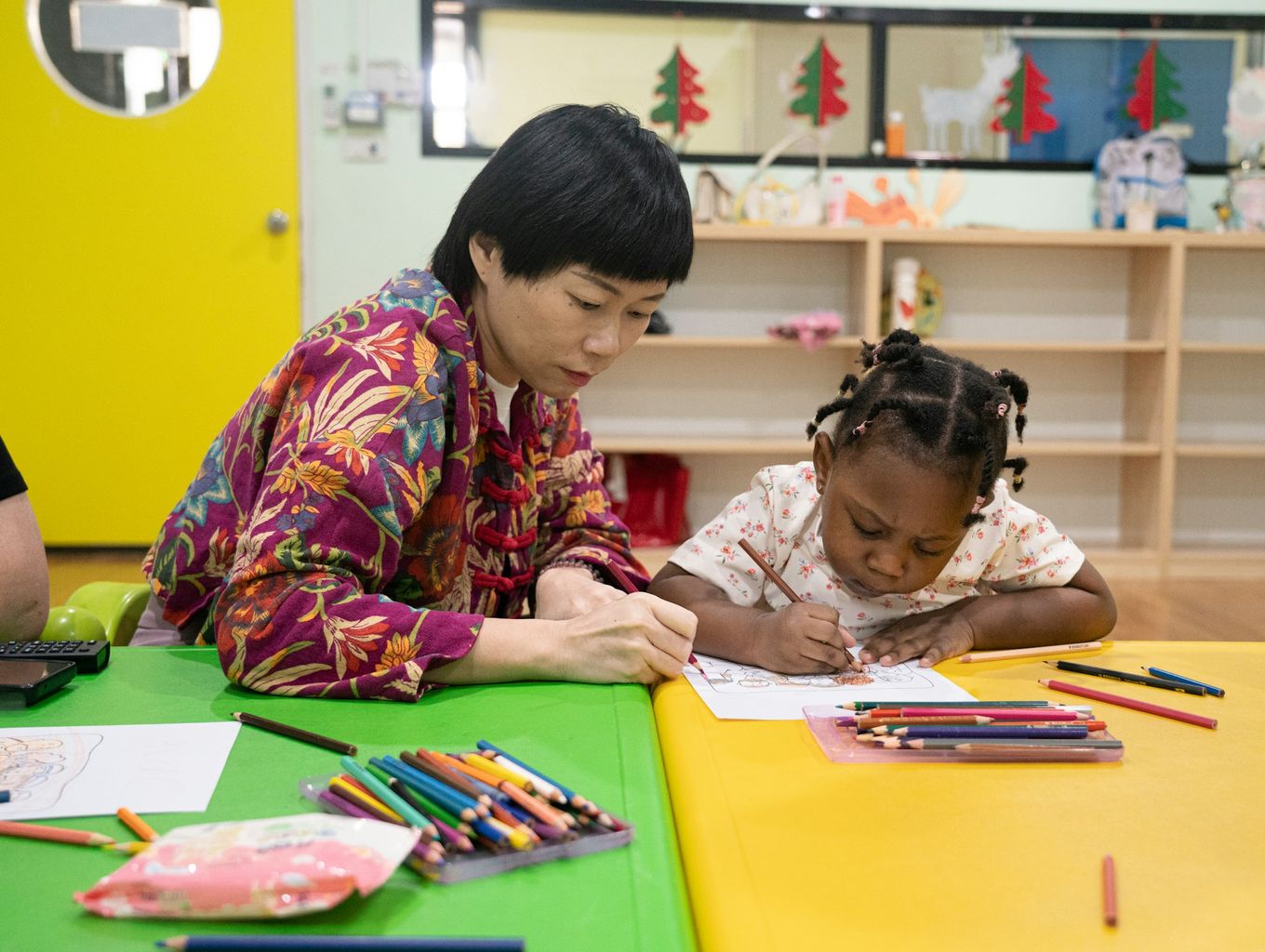 Teacher helping a young girl draw at a preschool table